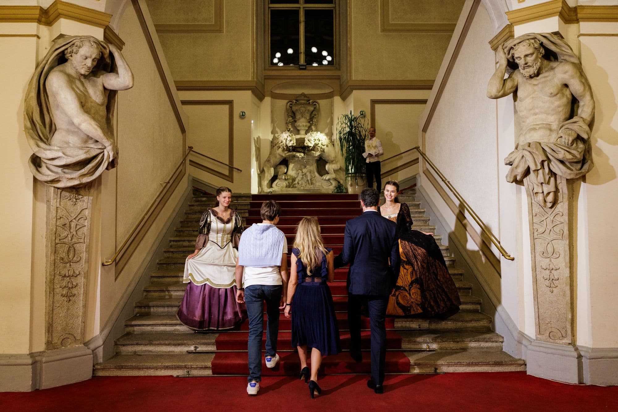 Guests climb the grand staircase with atlas statues, welcomed by hosts in historical costume