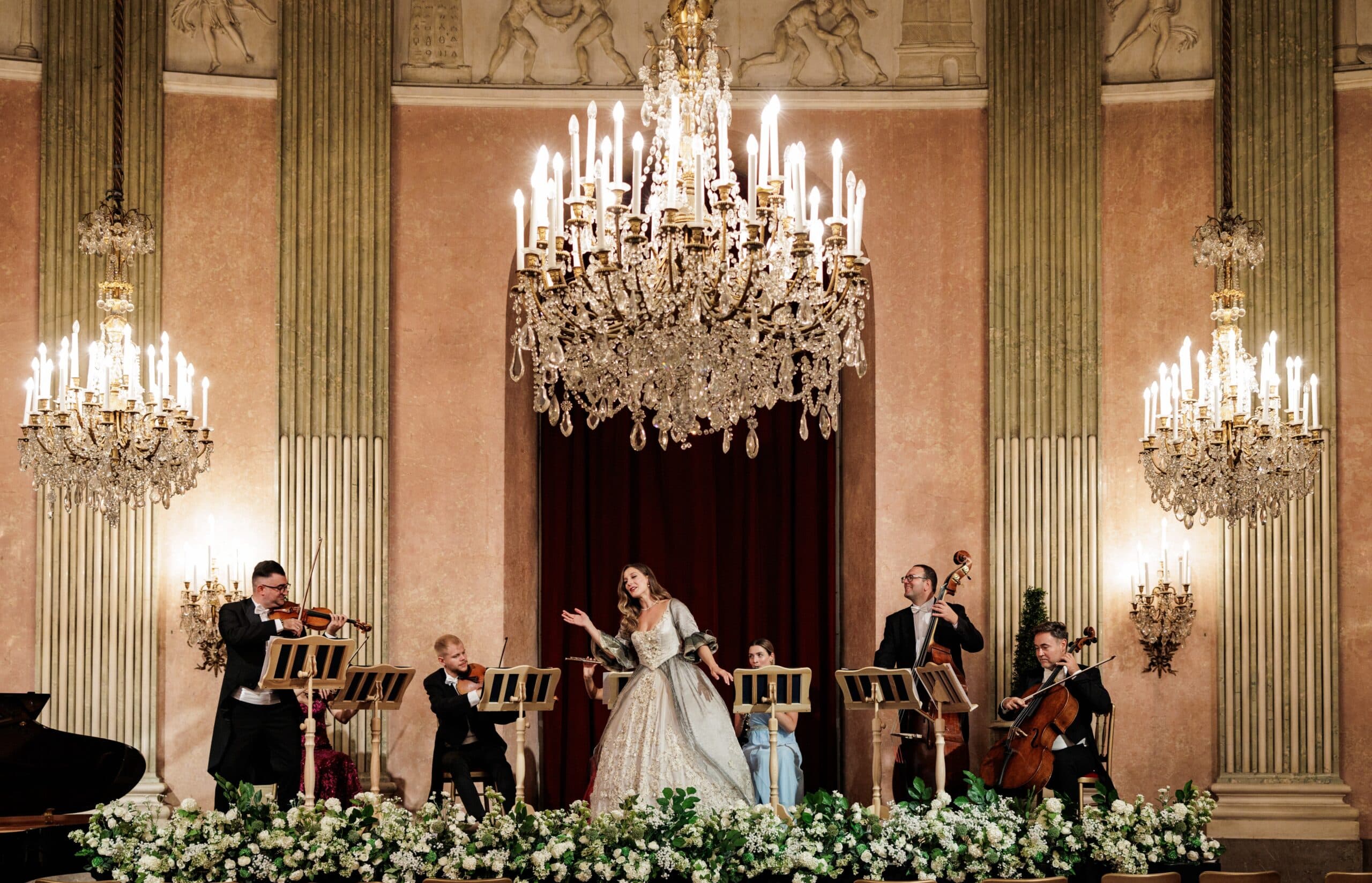Orchestra and soprano on a flower-lined stage under chandeliers