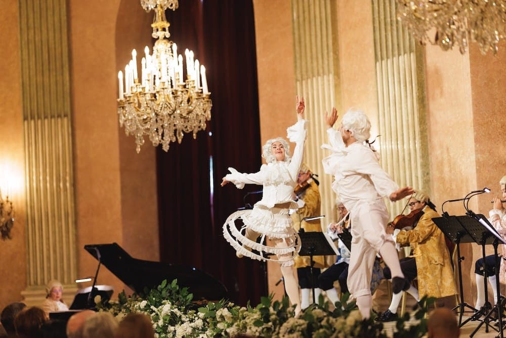 Two dancers in white rococo costumes perform in front of the orchestra