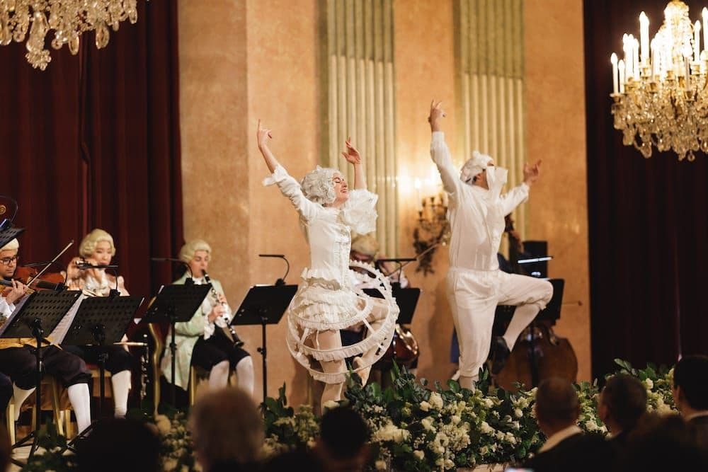Dancers in white rococo costumes leap in front of the audience