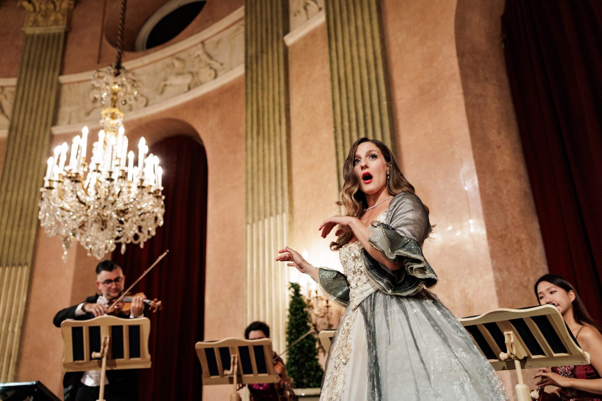 Soprano in a silver gown sings beside string players under a crystal chandelier