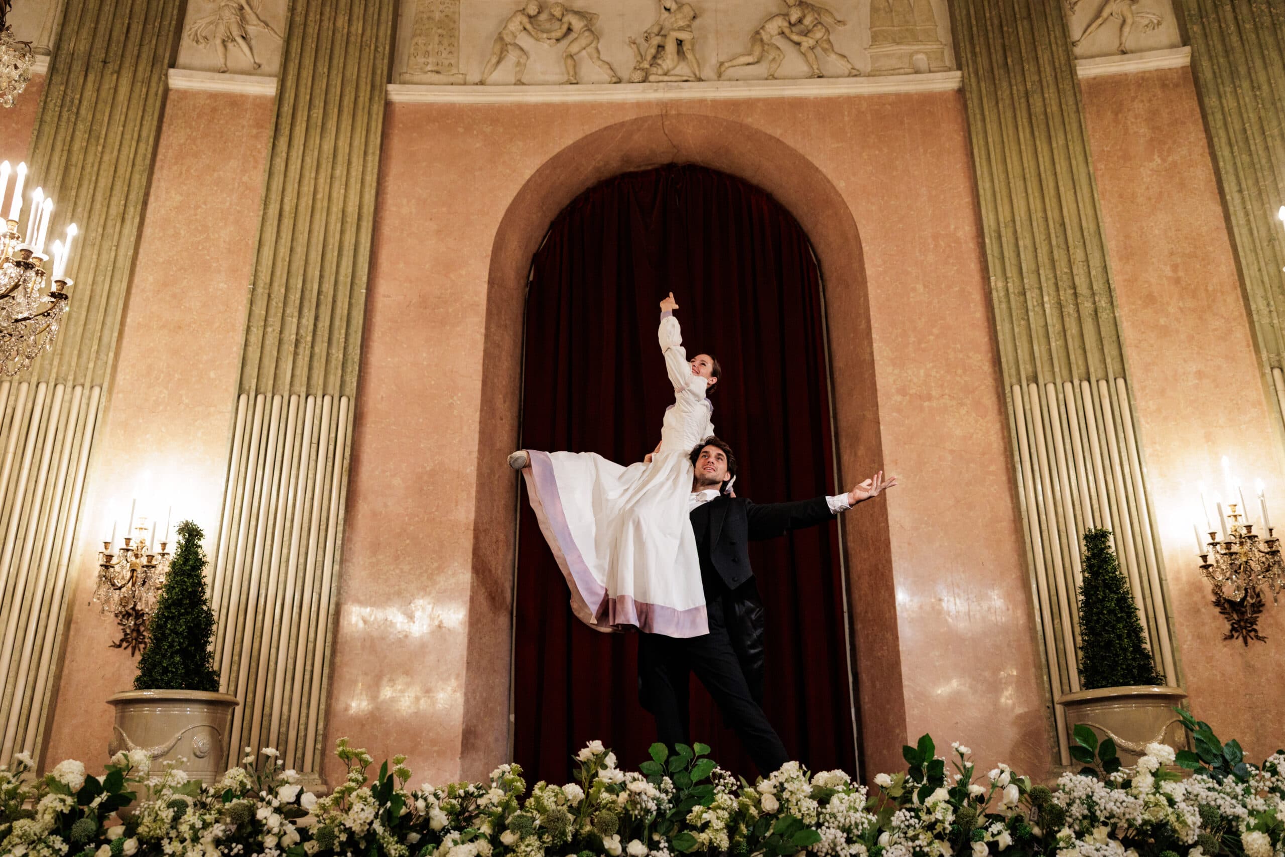Ballet couple in a lift beneath an arched doorway, white flowers in front