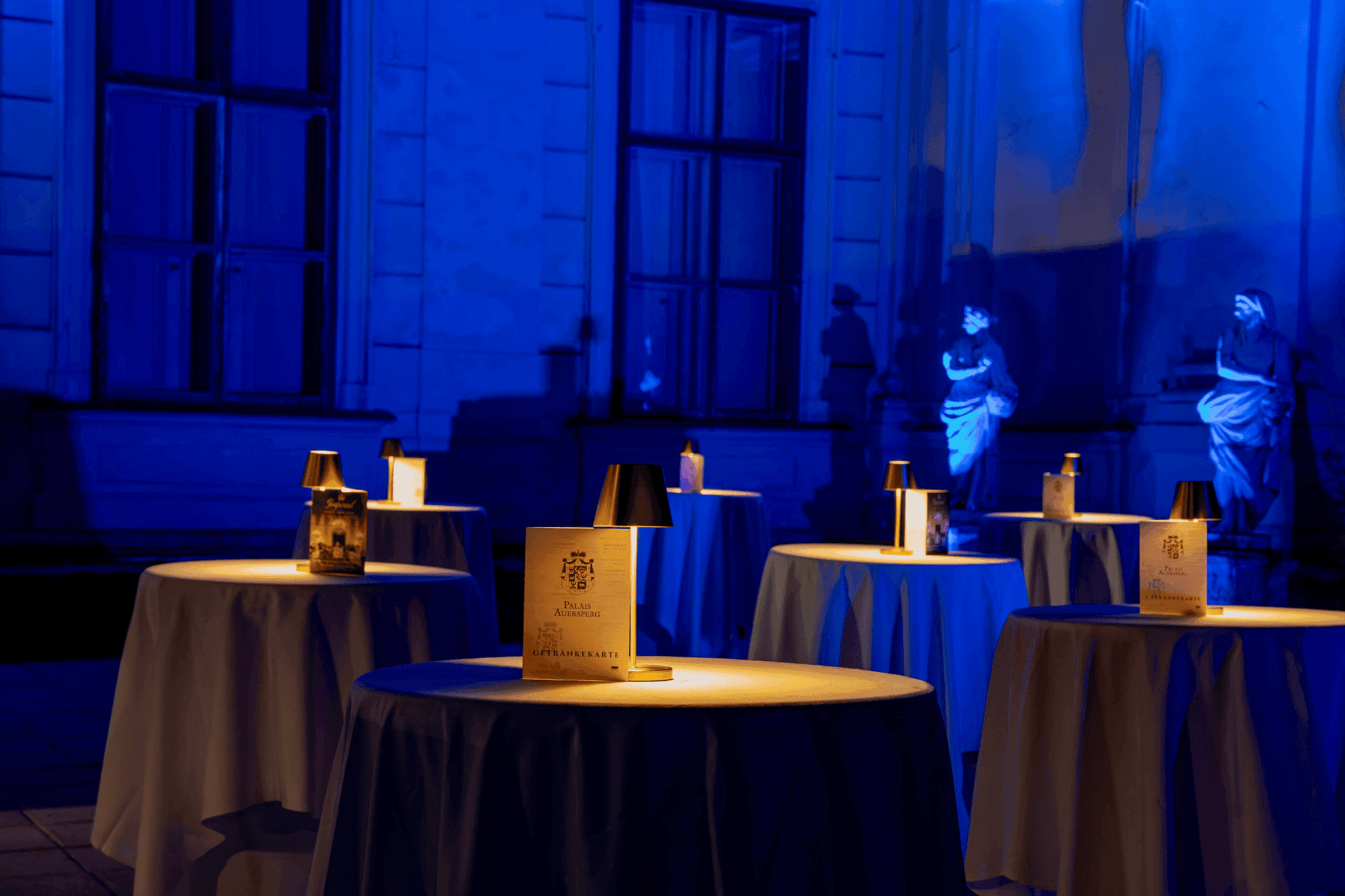 Courtyard at night in blue light with illuminated statues and cocktail tables