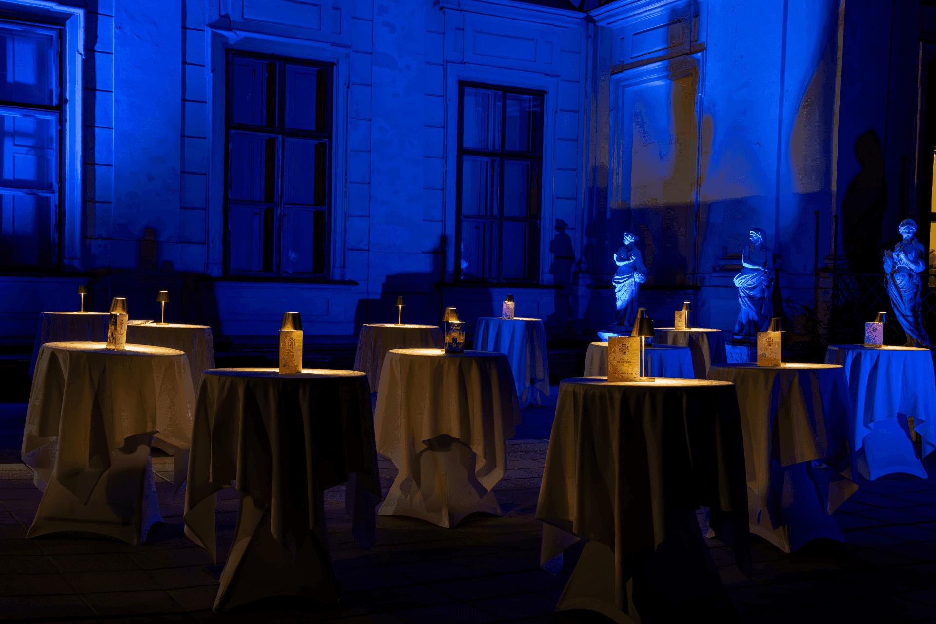 Courtyard at night in blue light with illuminated statues and cocktail tables