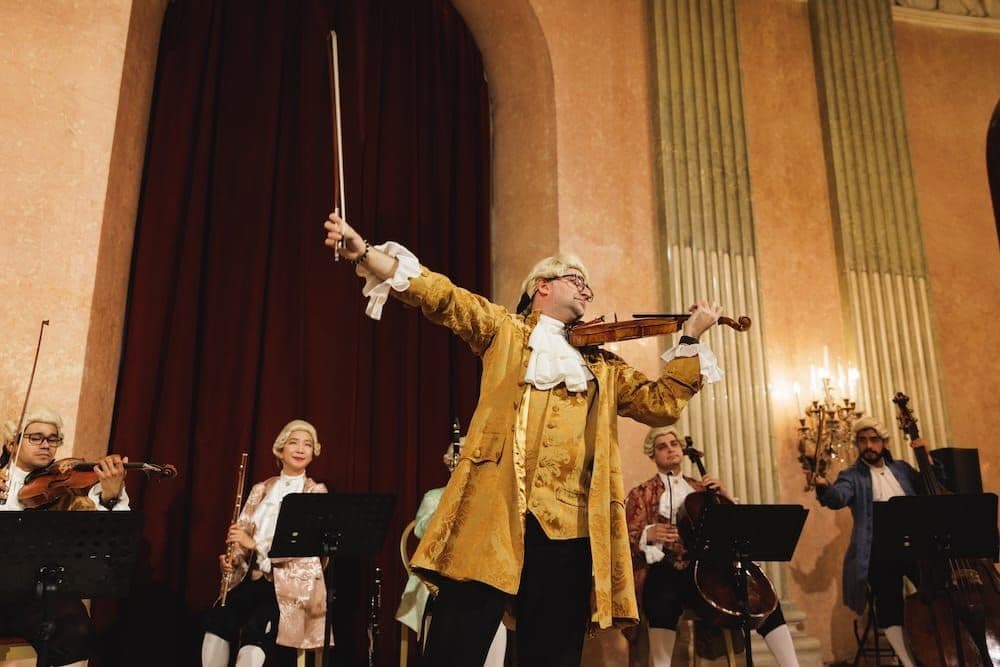 Violinist in a gold Mozart-era costume raises his bow before the orchestra