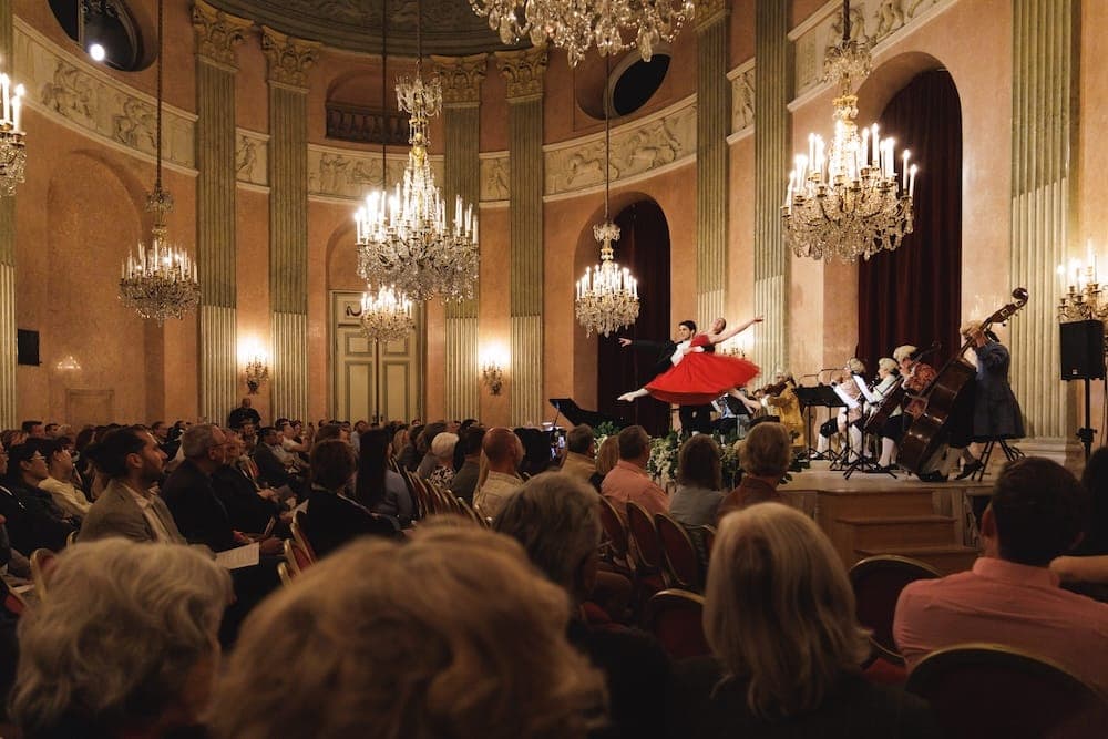 Packed hall under chandeliers as a dancer in red performs with the orchestra