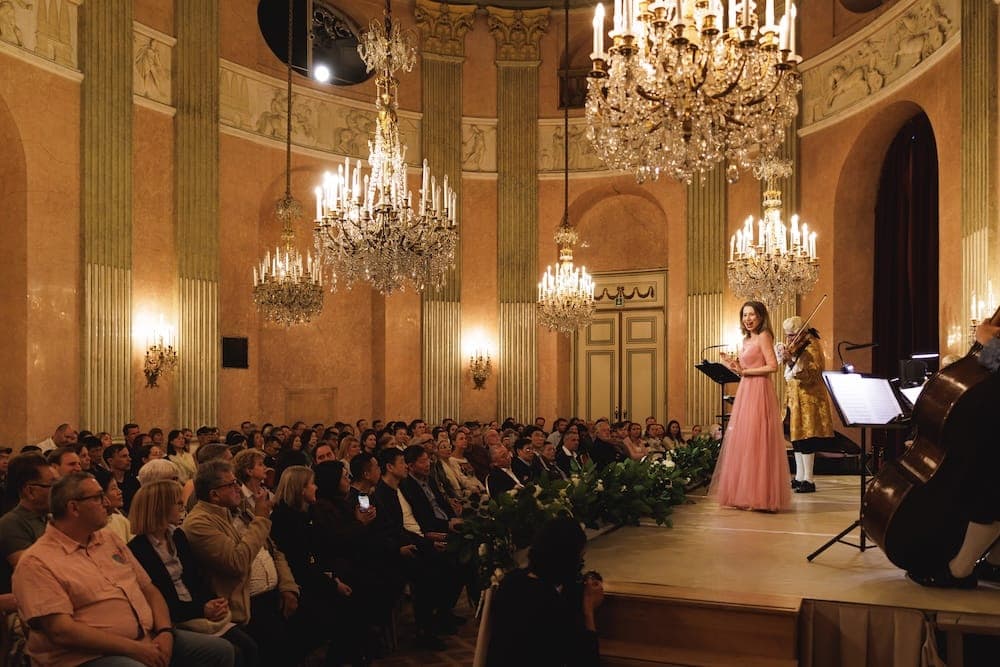 Singer in a pink gown on stage before a full audience under chandeliers